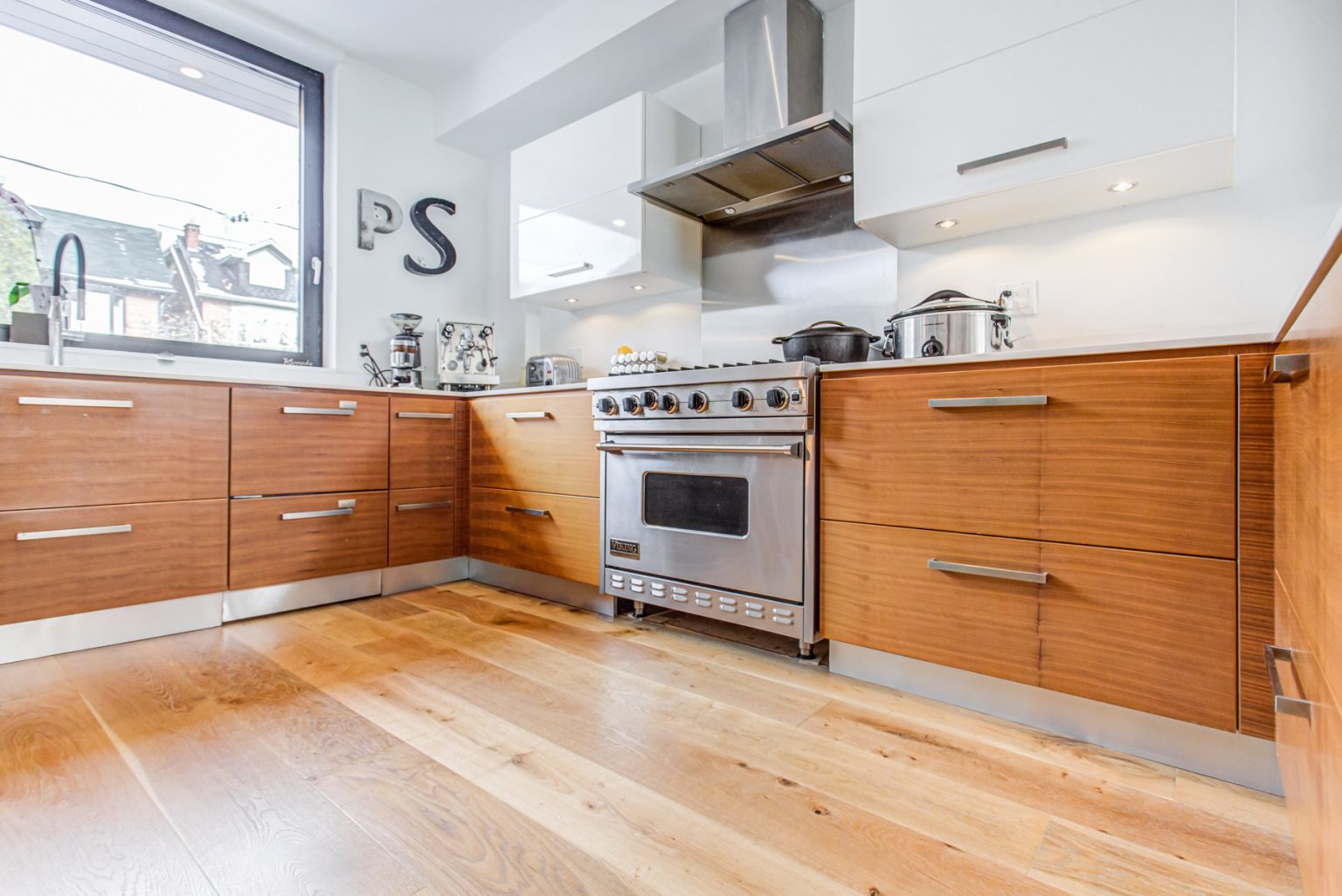 Modern kitchen equipped with artist retreat2 wooden cabinets, stainless steel appliances, and a light wooden floor. A large window provides natural light, while decorative "P" and "S" letters adorn the wall.
