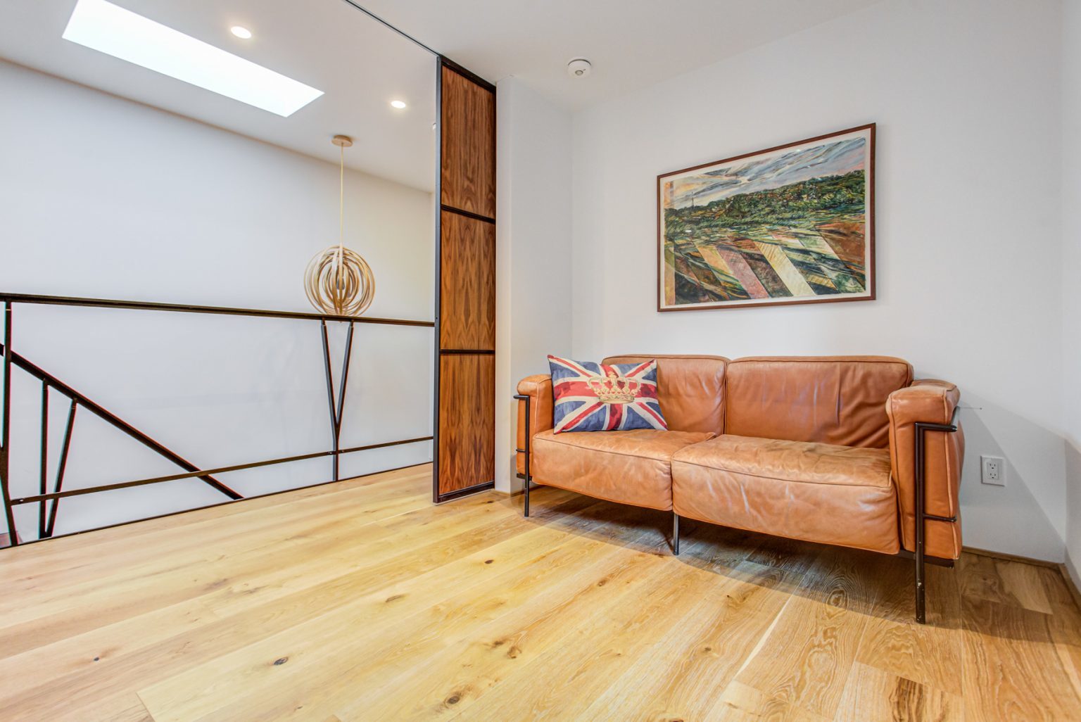 A modern interior space featuring a tan leather couch with a Union Jack pillow, hardwood floors, a framed landscape painting from the Artists Retreat 5 collection on the wall, and a decorative light fixture above a staircase.