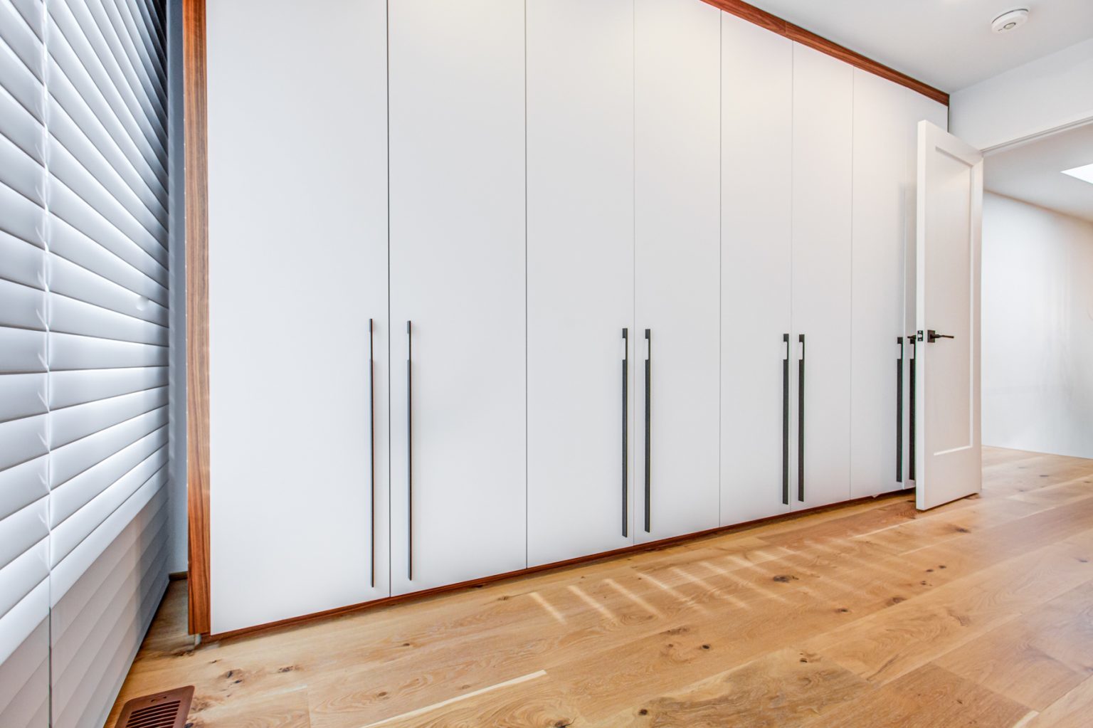 A row of closed artists retreat6 white wardrobe doors with long black handles stands in a bright room with wooden flooring. An adjacent door is partially open, and there are closed blinds on a window to the left.