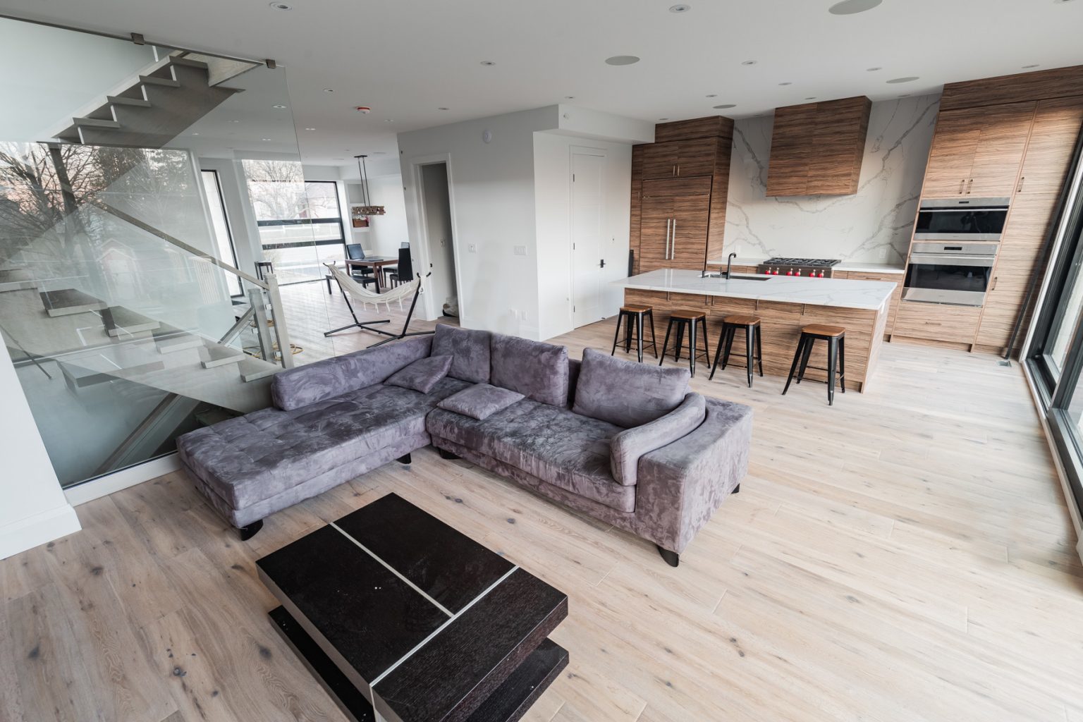 A modern open-concept living space featuring the Oak Oasis 1 sectional sofa in gray, a sleek black coffee table, a stylish kitchen island complete with bar stools, elegant wooden cabinets, and a sophisticated glass staircase. The room is bathed in natural light from large windows and showcases beautiful light wood flooring.