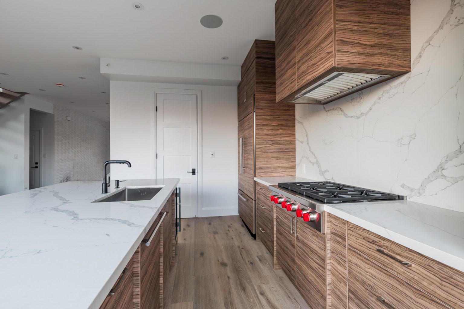 Modern kitchen with oak-finished cabinets (Oak Oasis 3), marble countertops, stainless steel gas stove with red knobs, and built-in appliances. The space features a sink with a black faucet on the island.