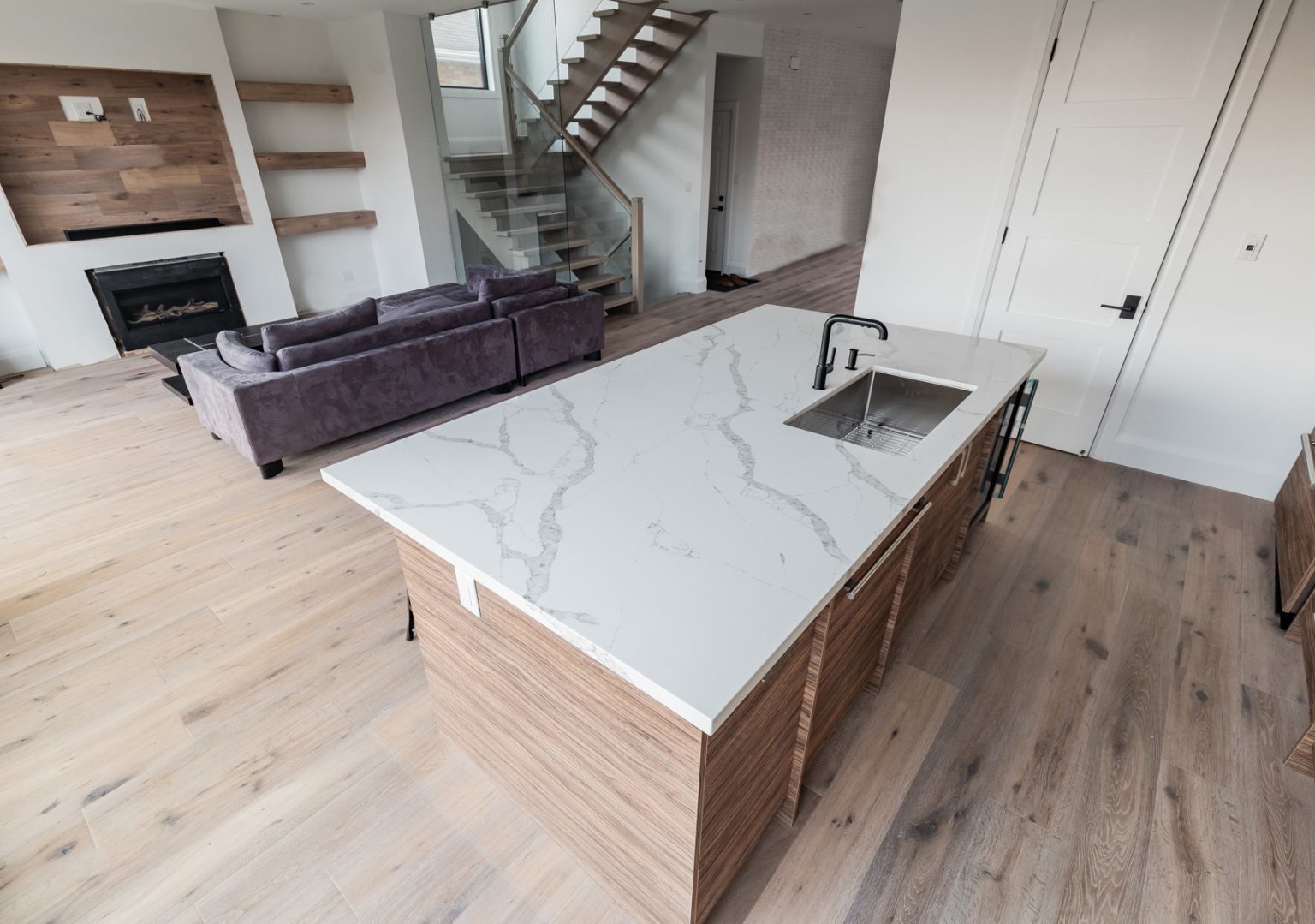 In a modern open-concept living space, the kitchen island boasts a sleek marble countertop. An inviting lavender sofa is positioned to enjoy the warmth of the fireplace on the left, while a stylish wooden staircase ascends to the upper floor, enhancing the room's aesthetics with its oak oasis 4 charm.